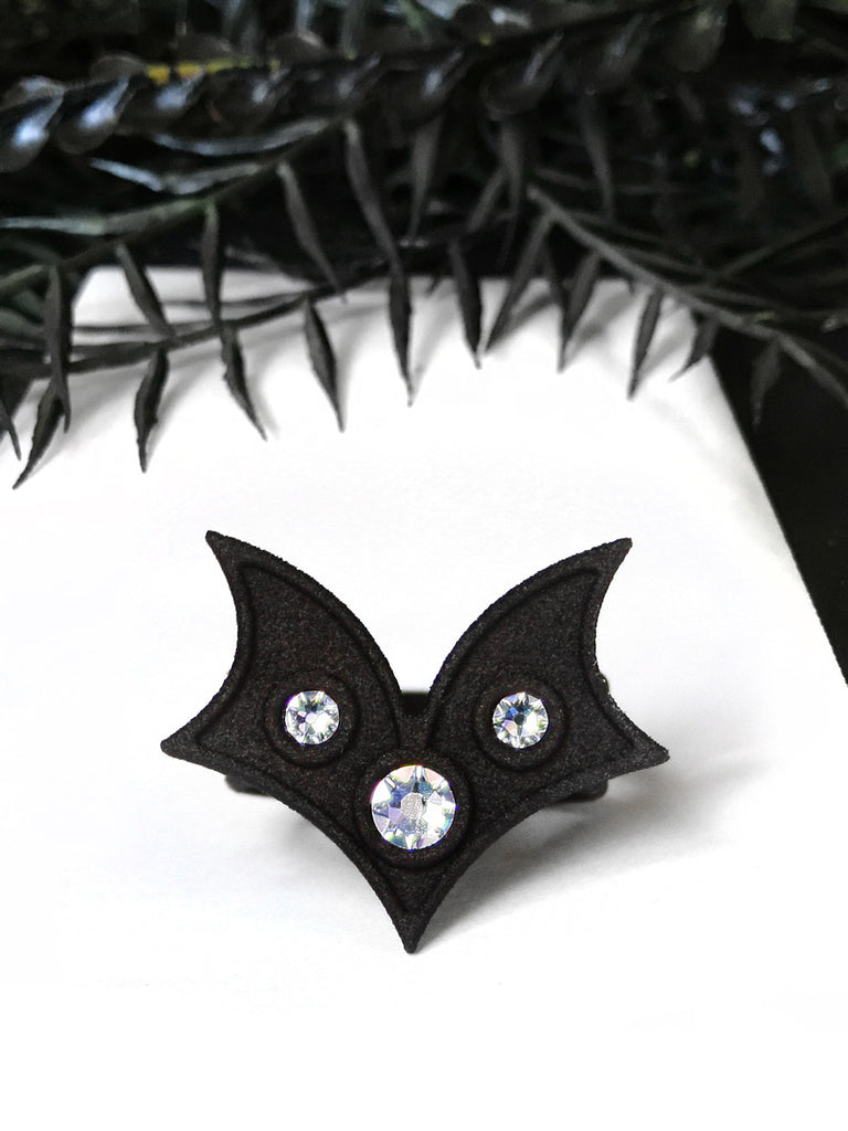 Matte black bat wing ring with white swarovski crystal stones, displayed on a white background with gothic black foliage. Witchy 3d printed jewelry by Hypnovamp.