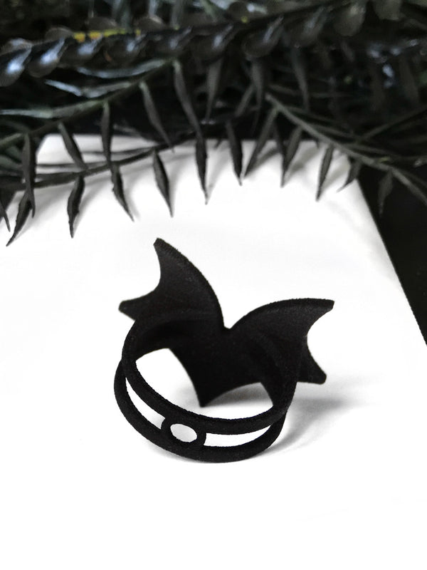 Back side view of matte black bat wing ring with white swarovski crystal stones and double bands, displayed on a white background with black foliage. Witchy 3d printed jewelry by Hypnovamp.