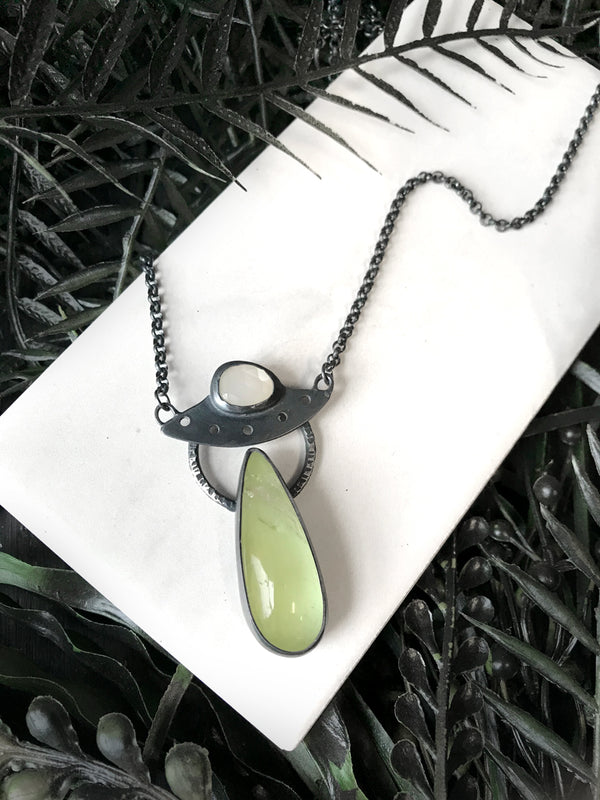 Handmade glow in the dark UFO necklace with moonstone and quartz, displayed on a white tile with black foliage. Inspired by the classic cult horror film Plan 9 from Outer Space. Created by Salem MA jewelry designer Hypnovamp.