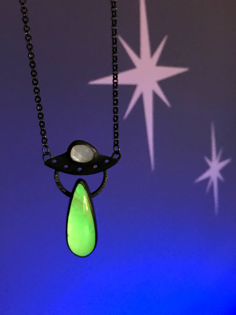 Handmade glow in the dark UFO necklace with moonstone and quartz, displayed against a blue background with retro atomic starbursts. Inspired by the classic cult horror film Plan 9 from Outer Space. Created by Salem MA jewelry designer Hypnovamp.