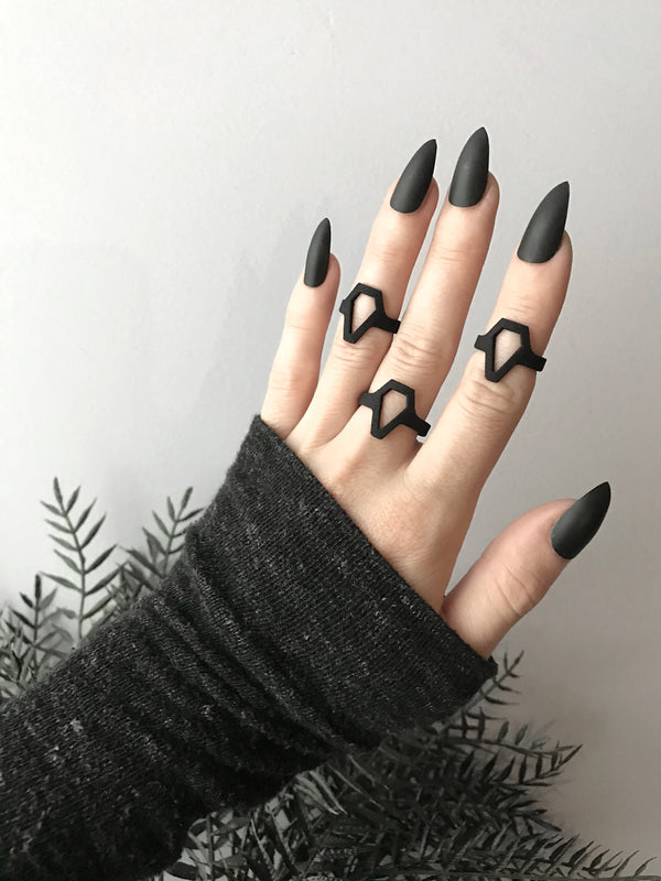Simple black coffin ring displayed on a gothic model hand with long black nails and witchy black foliage. Minimalist 3d printed jewelry by Hypnovamp.