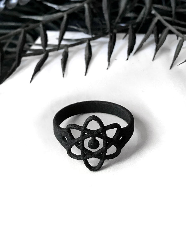 Matte black atomic symbol ring on a white background with witchy black foliage. Gothic 3d printed jewelry by Hypnovamp.