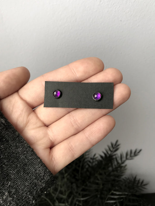 Witchy style stud earrings featuring oxidized sterling silver & rose-cut purple amethyst. Displayed in hand with black foliage in the background. Minimalist gothic jewelry handmade in Salem, MA by jewelry designer Hypnovamp.