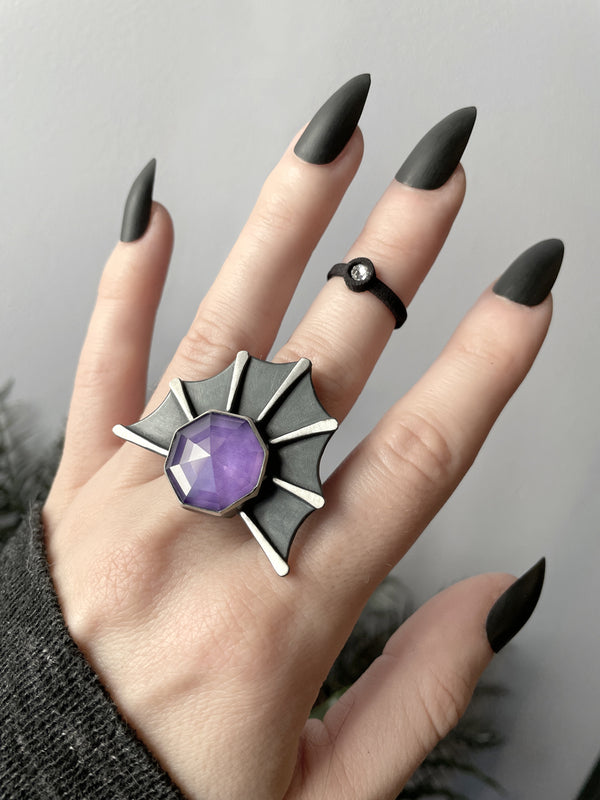 Handmade gothic bat wing ring with pink gemstone in the center, worn on a witchy model hand with long black nails and a simple black ring. Handmade silver glow in the dark jewelry by Salem MA artist Hypnovamp