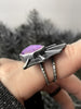 Side view of gothic bat wing ring with light purple gemstone in the center, worn on a hand with a simple black ring. Handmade silver glow in the dark jewelry by Salem MA artist Hypnovamp