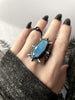 Blue glowing gemstone ring inspired by the Creature from the Black Lagoon, with sterling silver bat fins, displayed on a gothic model hand with long black nails and classic black ring, with black foliage in the background. Handmade witchy silver glow in the dark jewelry by Salem, MA artist Hypnovamp.