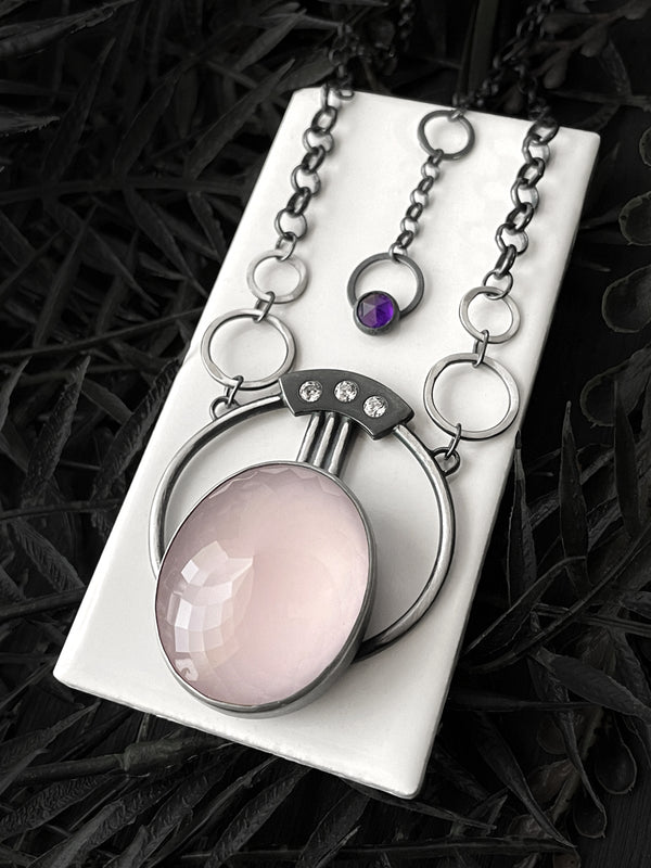 Handmade silver space-age art deco necklace with faceted rose quartz and amethyst, displayed on a white tile against a floral black background. Fancy glow in the dark jewelry made by Salem, Massachusetts artist Hypnovamp.