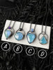 Blue ice zircon crystal glow necklaces with amethyst, black spinel, white and rainbow moonstone, displayed on a white tile again a black floral background and the variant letters A B C and D. Handmade silver glow jewelry by Salem, Massachusetts artist Hypnovamp. 