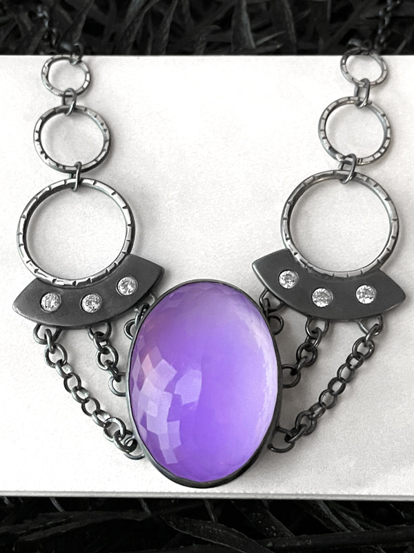Space age purple glow in the dark necklace with a large rose quartz crystal ball and silver UFOs with chain and crystals, displayed on a white tile with black foliage in the background. Handmade silver glow jewelry by Salem MA artist Hypnovamp