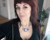 Portrait of Stephanie Smiszek, the artist behind Hypnovamp jewelry. She has dark red hair and pale skin, is wearing black spiderweb earrings and an art deco rose quartz necklace.