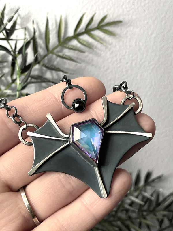 Handmade silver bat necklace with purple/blue glowing gemstone, displayed in a hand with black foliage in the background. High quality gothic glow in the dark jewelry made in Salem Massachusetts by jewelry artist Hypnovamp.