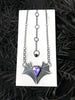 Handmade silver bat necklace with pink & purple gemstone, displayed on a white tile against black foliage in the background. Unusual gothic glow in the dark jewelry made in Salem MA by jewelry designer Hypnovamp.