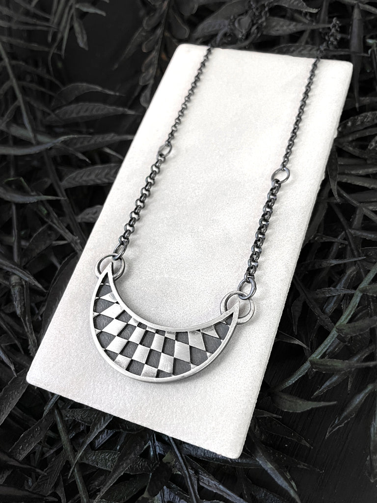 Warped checkerboard op art pattern inside a moon. Handmade silver necklace by Hypnovamp.