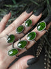 Green gemstone rings displayed on a gothic model hand with long matte black nails and dark foliage. Witchy chrome diopside jewelry by Salem Massachusetts jewelry designer, Hypnovamp.