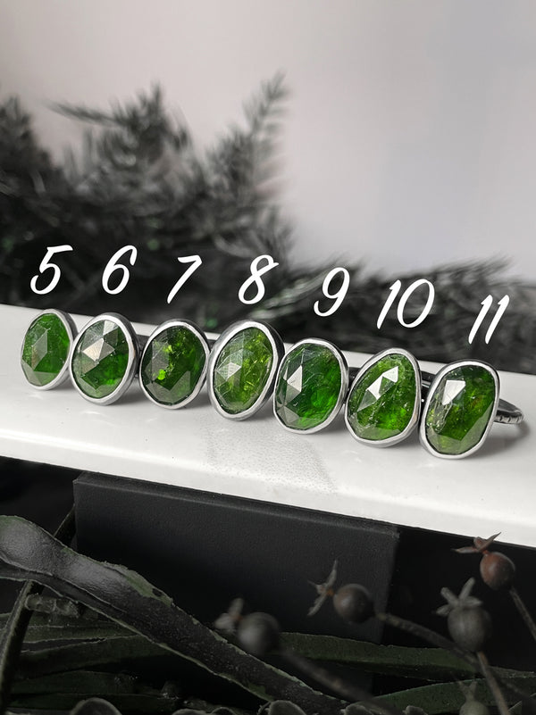 A row of sterling silver and vibrant green gemstone rings with the sizes 5-11 above them. Witchy chrome diopside jewelry creates by silversmith & jewelry designer Hypnovamp in Salem Massachusetts.