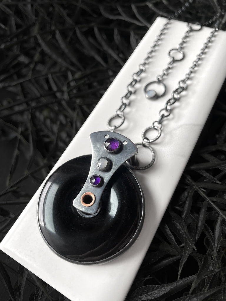 Handmade black onyx necklace with amethyst and moonstone, displayed on a white tile against black foliage. Kinetic spinning wheel of fortune jewelry handmade in Salem MA by jewelry designer Hypnovamp.