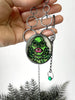 Handmade Silver and quartz necklace featuring a glow in the dark painting inspired by the creature from the black lagoon. Displayed in a hand with black foliage in the background. Horror and glow jewelry by Salem, MA artist Hypnovamp.