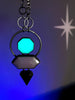 Night time view of space witchy statement glowing necklace with art deco style & 80s mall colors, displayed on a white tile against black foliage.. High quality handmade glow in the dark jewelry by Hypnovamp.