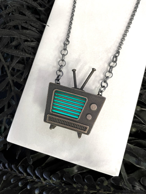 Vintage mid century television set necklace with glow in the dark blue static screen, displayed on a white tile with black leaves in the background. Spooky retro glow jewelry created by Salem, MA artist Hypnovamp.