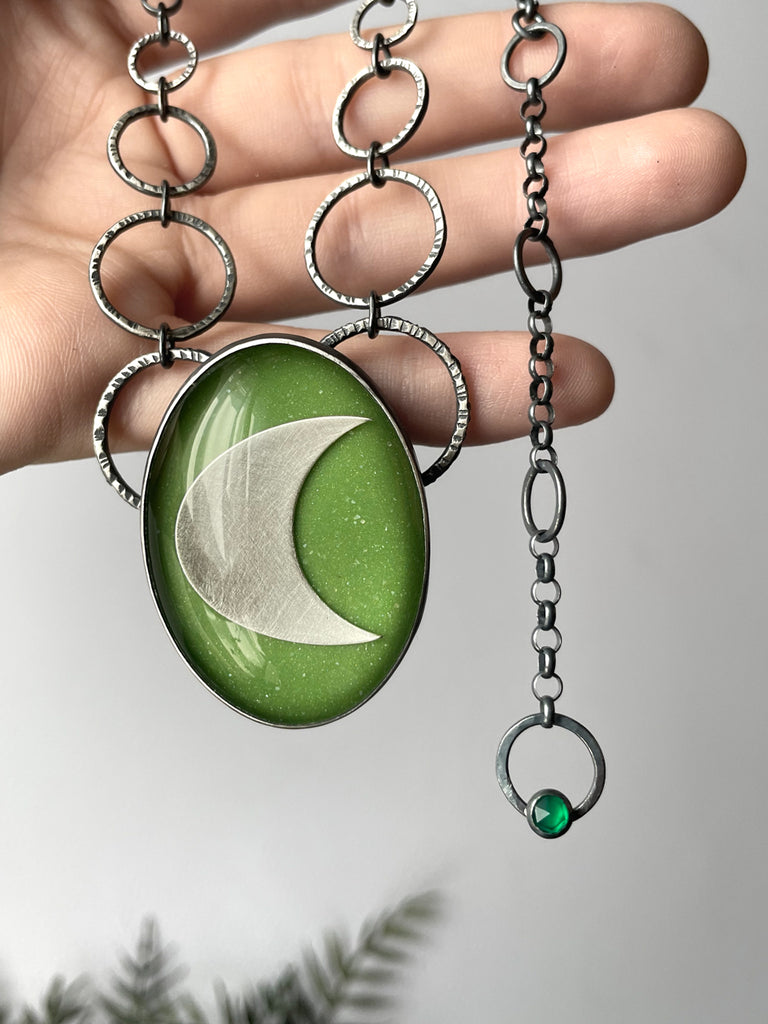 Handmade silver glow necklace with a witchy sci-fi space vibe. Featuring glow in the dark green glass with a silver crescent moon and tiny green onyx. Displayed in a hand. High quality glow jewelry by Salem, MA artist Hypnovamp.