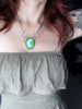 Handmade silver glow necklace with a witchy sci-fi space vibe. Featuring glow in the dark green glass with a silver crescent moon and tiny green onyx. Displayed on tattooed model wearing a brown dress. High quality glow jewelry by Salem, MA artist Hypnovamp.