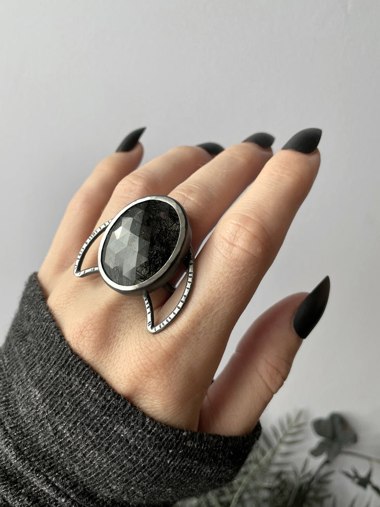 Witchy black rutilated quartz ring with a crescent moon shape, displayed on a gothic model's hand with long black nails. Sterling silver glow jewelry handmade in Salem, MA by artist Hypnovamp.