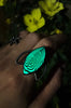 Night view of handmade silver glow ring with trippy op art pattern under a marquise shaped stone, set on a silver crescent moon. Displayed on a gothic model hand with yellow flowers. High quality glow in the dark jewelry made in Salem, MA by artist Hypnovamp.