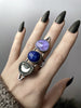 Space queen statement ring with blue lapis lazuli, glowing purple rutilated quartz, & clear quartz with a silver moon, displayed on a witchy model's hand with long black nails. High quality handmade silver glow in the dark jewelry by Hypnovamp.