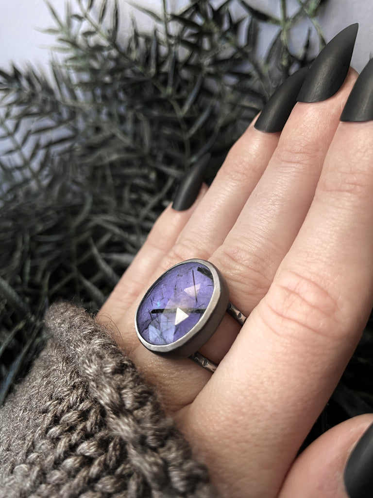 Handmade silver ring with purple glow in the dark gemstone (rutilated quartz) displayed on a gothic model hand with long black nails.