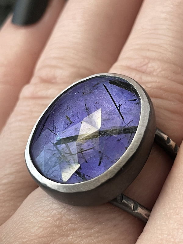 Close up of handmade silver ring with purple glow in the dark gemstone (rutilated quartz) displayed on a gothic model hand with long black nails.